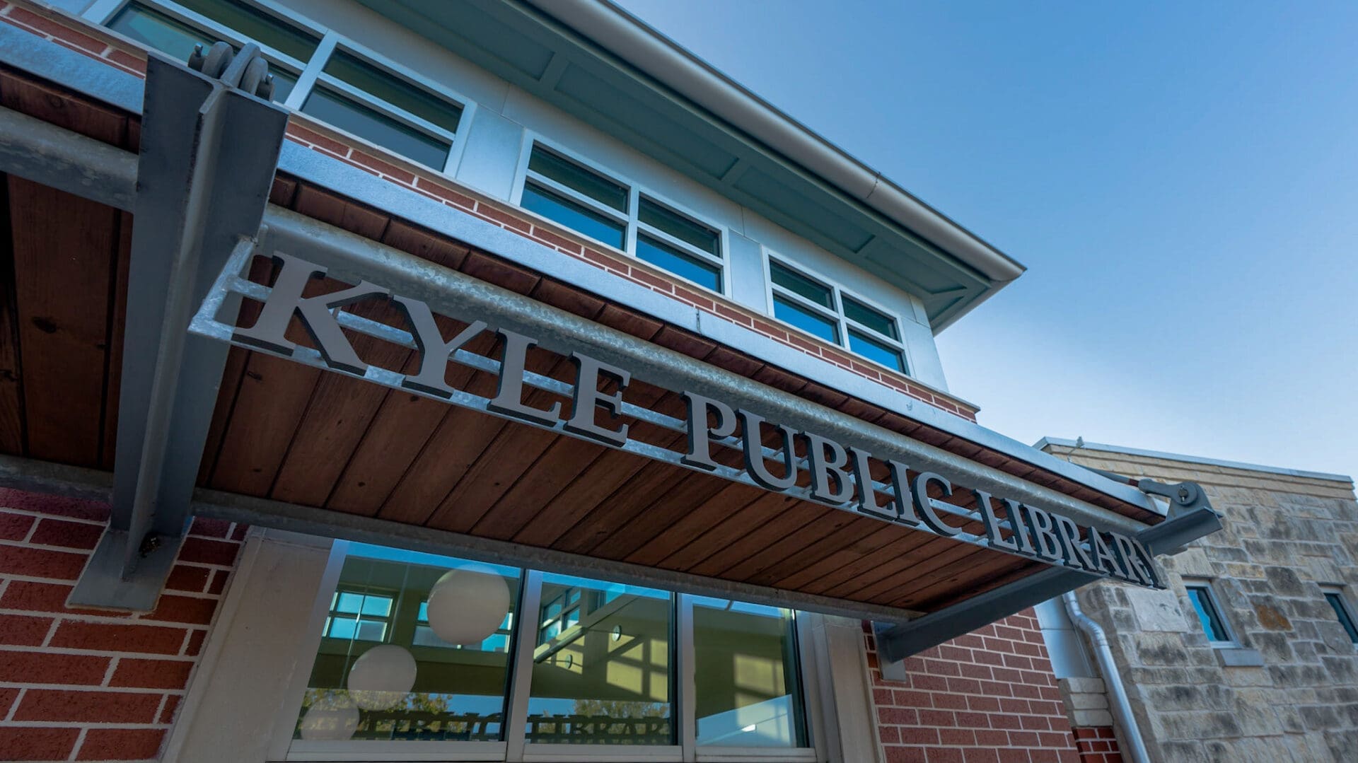 The image shows the exterior of a building with a sign that reads "Kyle Public Library" above the entrance, featuring brick and stone walls, large windows, and a blue sky in the background.