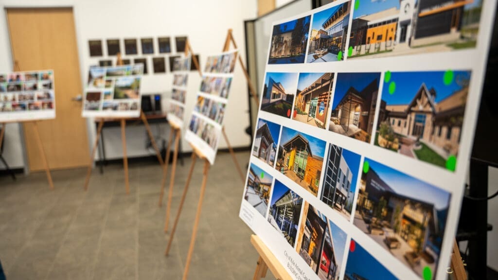 Photo of the Animal Care Center Public Input meeting in March 2025. A display of architectural design boards on easels in a room, each board featuring various building images and some colored stickers, with a door and plaques visible in the background.