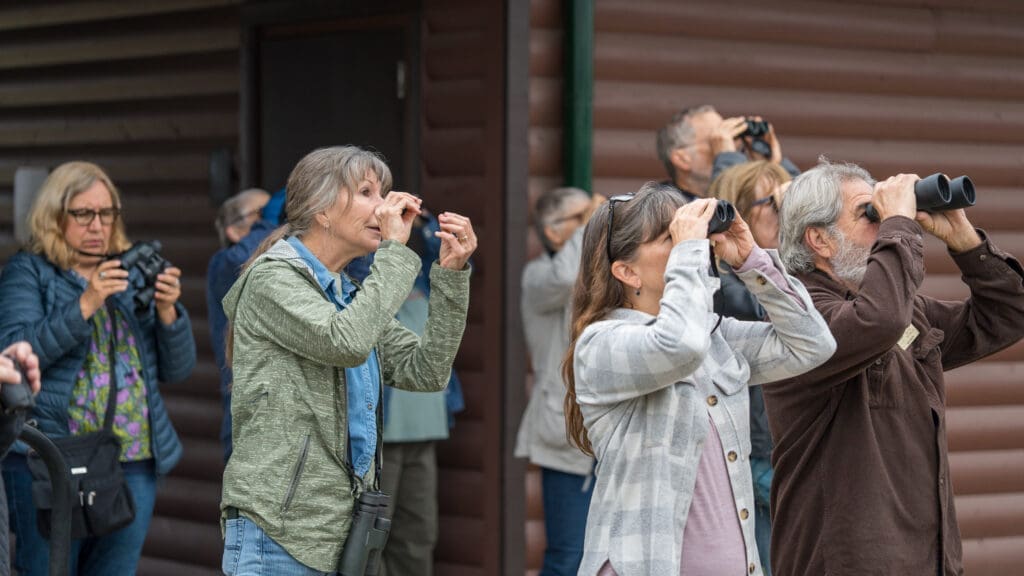 A group of adults stands outside a wooden building, looking up and using binoculars and cameras, possibly birdwatching or observing something in the sky at a Bird Walk at Lake Kyle. 