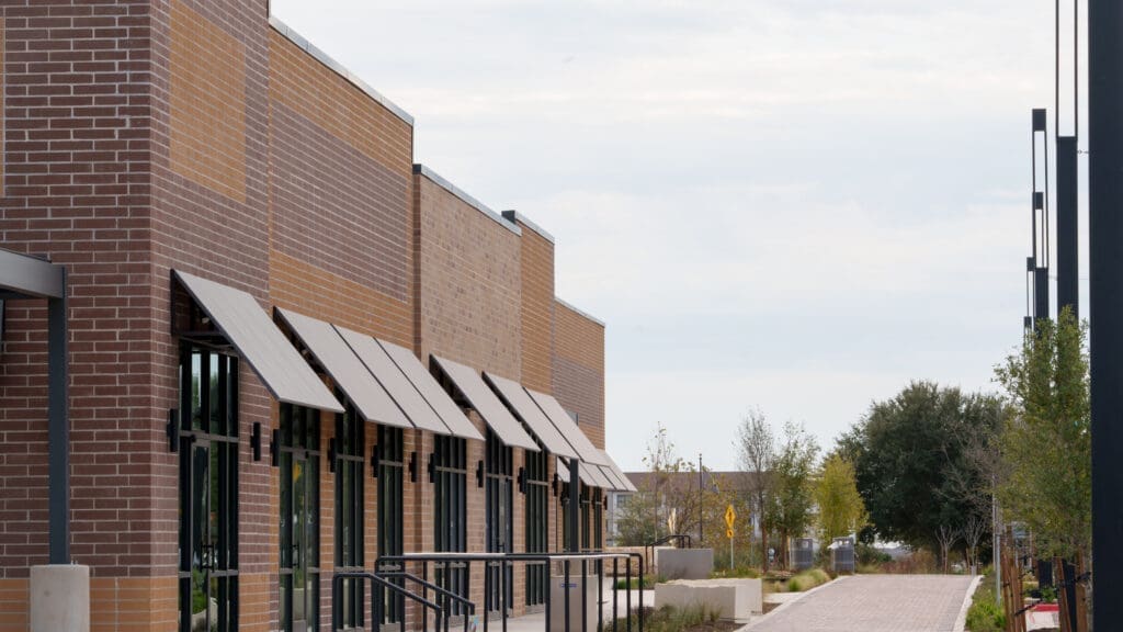 A row of modern, empty brick storefronts with large windows and metal awnings, situated along a landscaped walkway with trees and shrubs on a cloudy day. Located in the Brick and Mortar District near Sprouts Farmers Market along FM 1626.