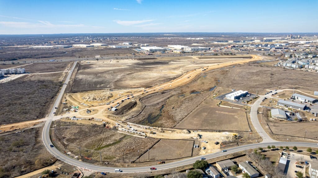 Aerial view of a construction site with dirt roads winding through a cleared area, surrounded by industrial buildings, roads, and undeveloped land under a clear blue sky.