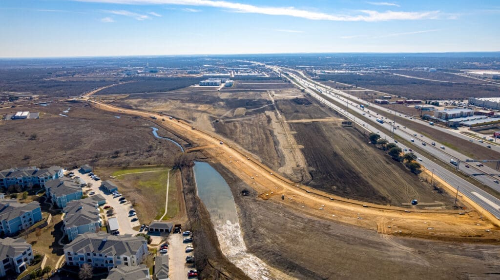 Aerial view of a highway expansion project beside a residential area, with cleared land, construction vehicles, a temporary pond, and nearby roads stretching into the distance under a blue sky.