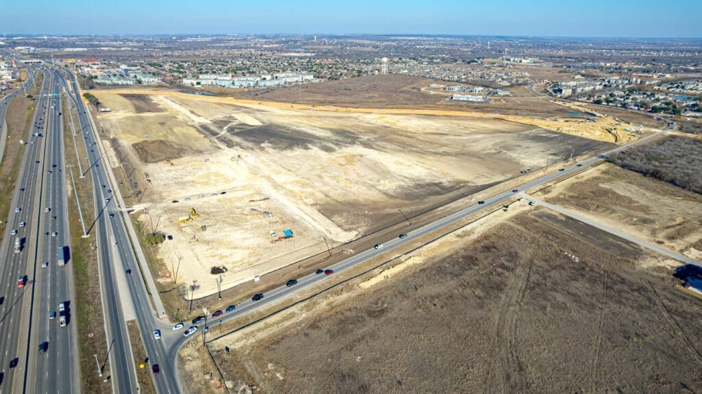 Aerial view of a large construction site next to busy highways, with cars driving by and developed city areas visible in the background. The land is mostly cleared and brown, with some equipment present.