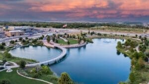 A curved pedestrian bridge crosses a peaceful pond surrounded by greenery, with an American flag, buildings, and a highway in the background under a colorful sunset sky. Drone image of Heroes Memorial Park in Kyle, TX.
