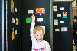 A smiling young girl points excitedly at her artwork displayed on a black panel with other children’s art pieces at an exhibit.