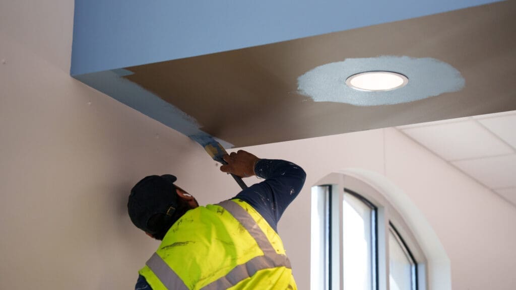 A worker in a yellow safety vest paints a ceiling edge blue, using a wide brush. There is a partially painted area around a recessed ceiling light nearby. Daylight comes through an arched window in the background.