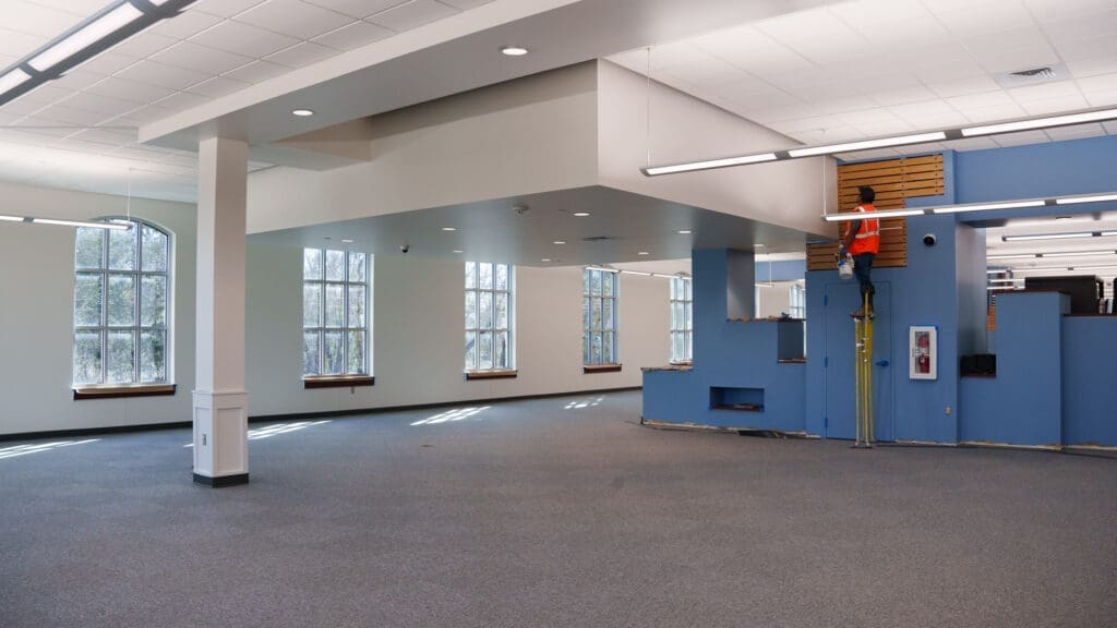 A worker in an orange vest stands on a raised blue platform inside a large, mostly empty room with carpeted floors, white walls, and large windows letting in natural light.