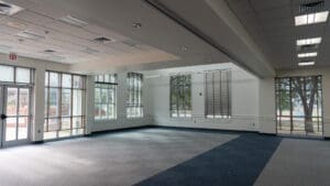 A large, empty room with carpeted floors, large windows with blinds, and overhead ceiling lights. Sunlight streams through the windows, and trees are visible outside. The space appears clean and modern.