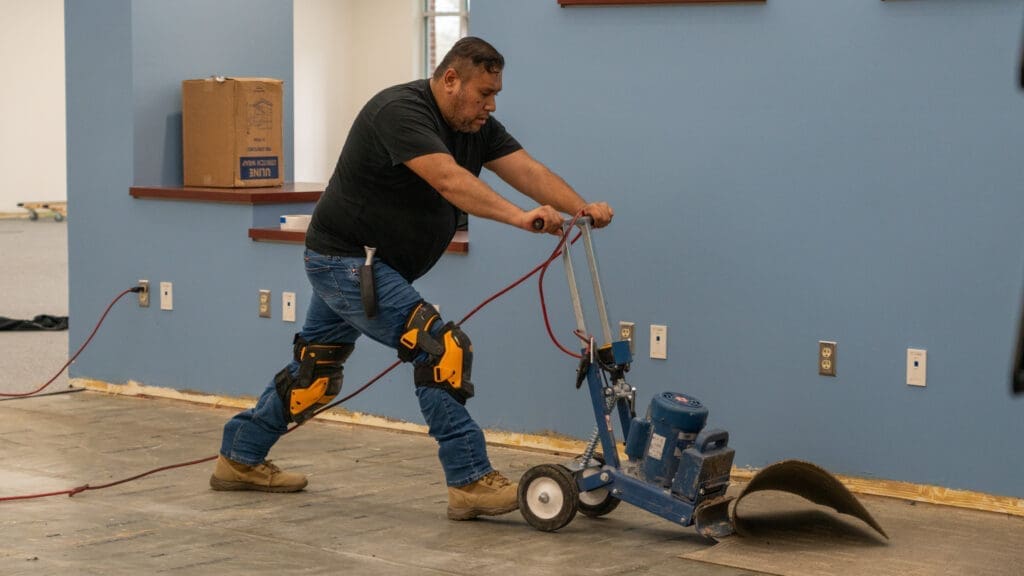 A man wearing knee pads uses a floor stripping machine to remove old flooring in a room with blue walls. A cardboard box sits on a wall shelf in the background.