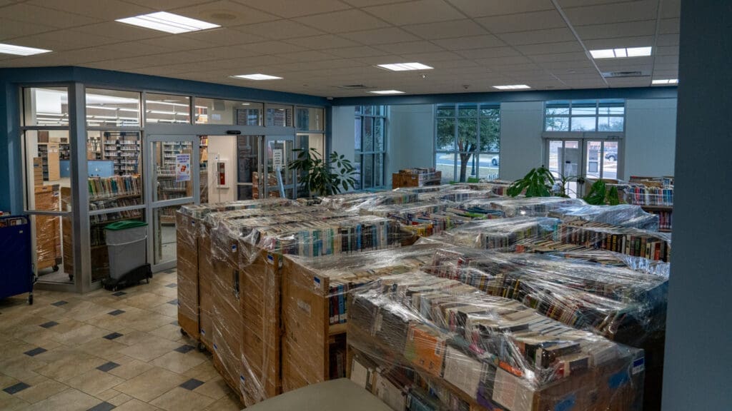 Rows of book carts wrapped in plastic are stacked in a library lobby near large windows and glass-walled offices. The area appears organized, with plants placed around the space.