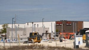 A construction site with heavy machinery and building materials in the foreground, and a Snooze restaurant with its logo on a brown and white building in the background. A chain-link fence surrounds the site. Future site of A Little Shady restaurant.
