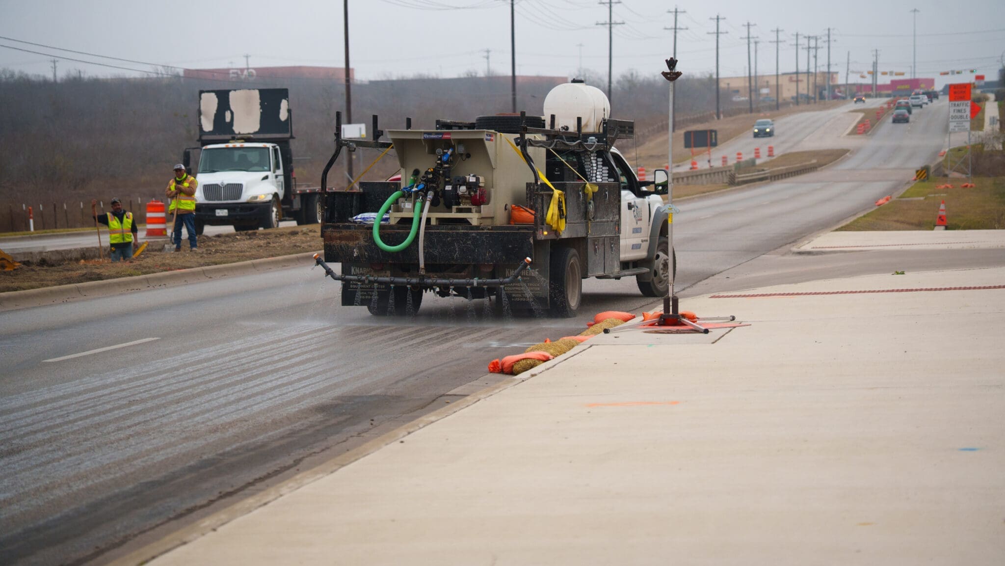 A City of Kyle truck sprays liquid on a road under construction while workers and construction equipment are visible in the background on a cloudy day. Traffic cones line the roadside.
