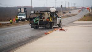 A City of Kyle truck sprays liquid on a road under construction while workers and construction equipment are visible in the background on a cloudy day. Traffic cones line the roadside.