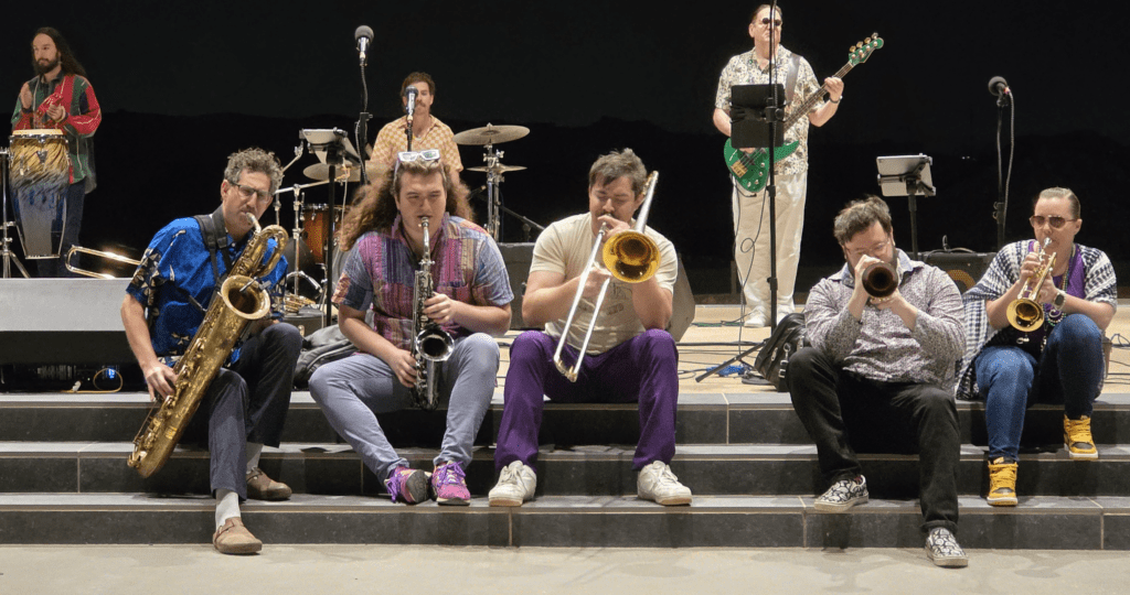 A band performs on stage: five musicians with brass instruments sit at the front, while four others play drums, congas, guitar, and keyboard in the background under stage lights at the Mardi Gras Celebration at La Verde Park.