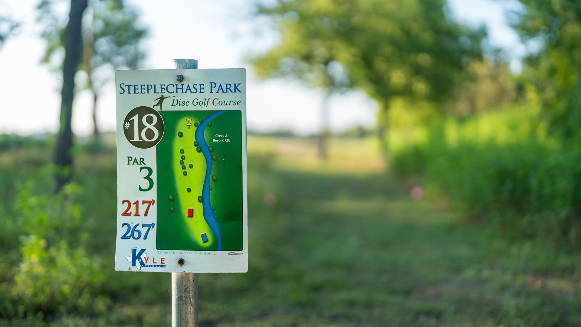A close-up of a Steeplechase Park disc golf course hole sign showing hole 18, par 3, with distances of 217 and 267 feet. The background features a grassy, tree-lined course under a clear sky.