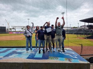 A group of seven people stands and celebrates on a platform near a baseball field. One person holds a certificate, and two people have their arms raised in excitement. The stadium and cloudy sky are visible in the background.