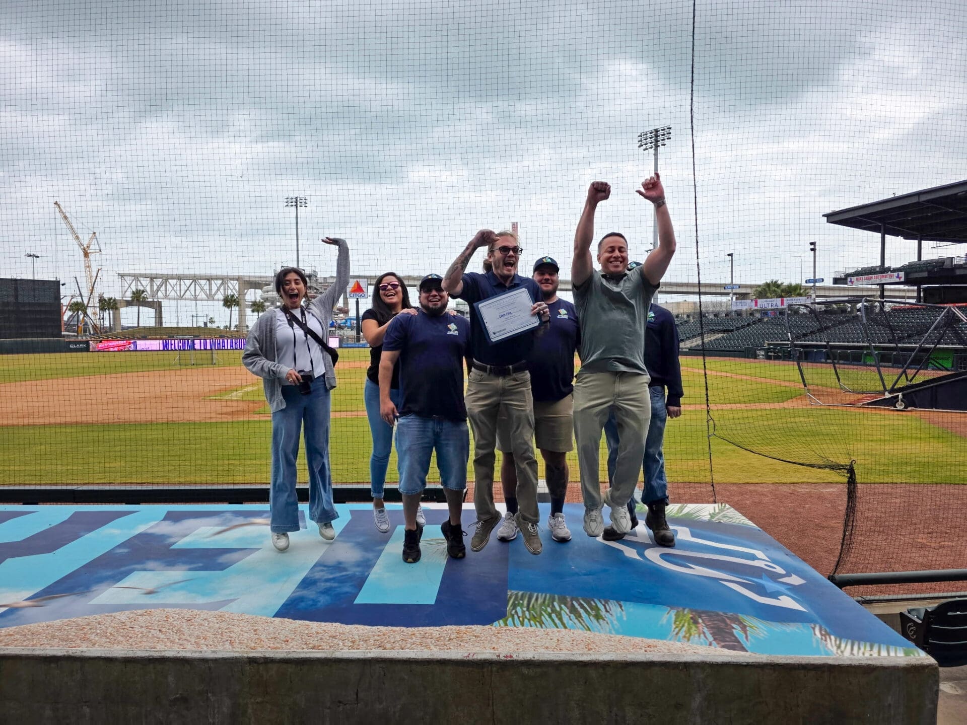 A group of seven people stands and celebrates on a platform near a baseball field. One person holds a certificate, and two people have their arms raised in excitement. The stadium and cloudy sky are visible in the background.
