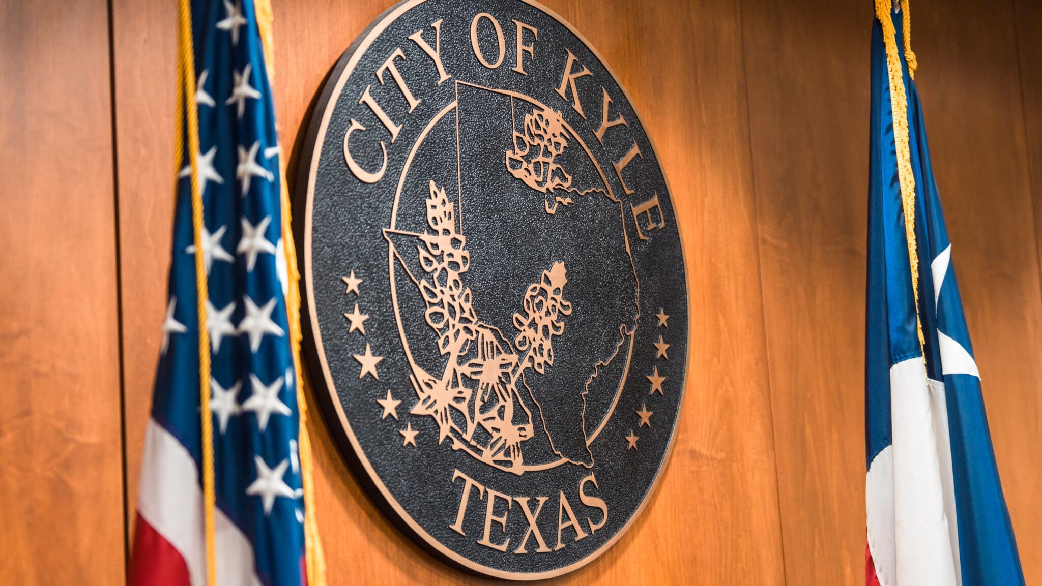 A wooden wall displays a large round seal reading "City of Kyle Texas" with a map outline of Texas. American and Texas flags are visible on either side of the seal.