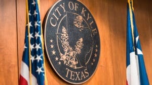 A wooden wall displays a large round seal reading "City of Kyle Texas" with a map outline of Texas. American and Texas flags are visible on either side of the seal.