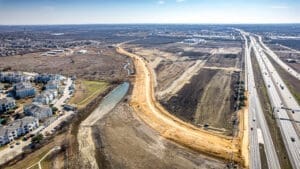Aerial view of a highway next to a large construction site with dirt roads, nearby pond, residential buildings to the left, and open undeveloped land in the background under a clear sky.