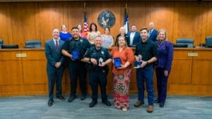 A group of people, including police officers holding plaques and civilians, pose for a photo in a council chamber with a city seal, American and Texas flags, and wood paneling in the background.