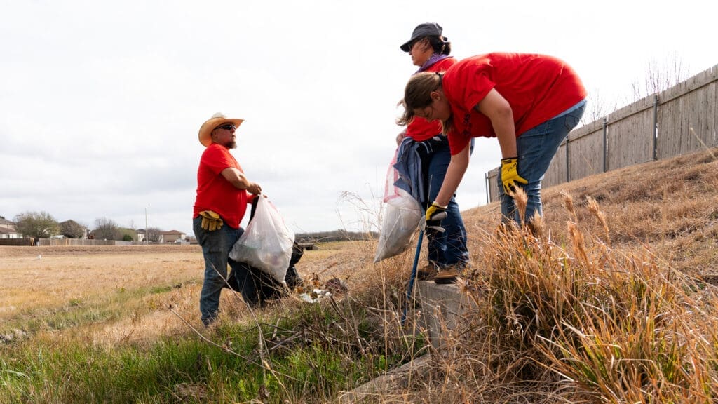 Three people in red shirts pick up trash and litter in a grassy, sloped field near a wooden fence. Two hold garbage bags, and one bends down collecting debris, all wearing gloves and jeans.