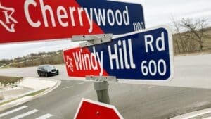 A close-up of intersecting street signs for Cherrywood and Windy Hill Rd at a street corner, with a black car approaching on the road and a cloudy sky in the background.