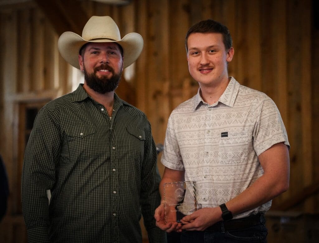 Two men stand indoors, smiling at the camera. One wears a cowboy hat and green plaid shirt, while the other, in a light patterned shirt, holds a clear glass award. The wooden wall behind them suggests a rustic setting.