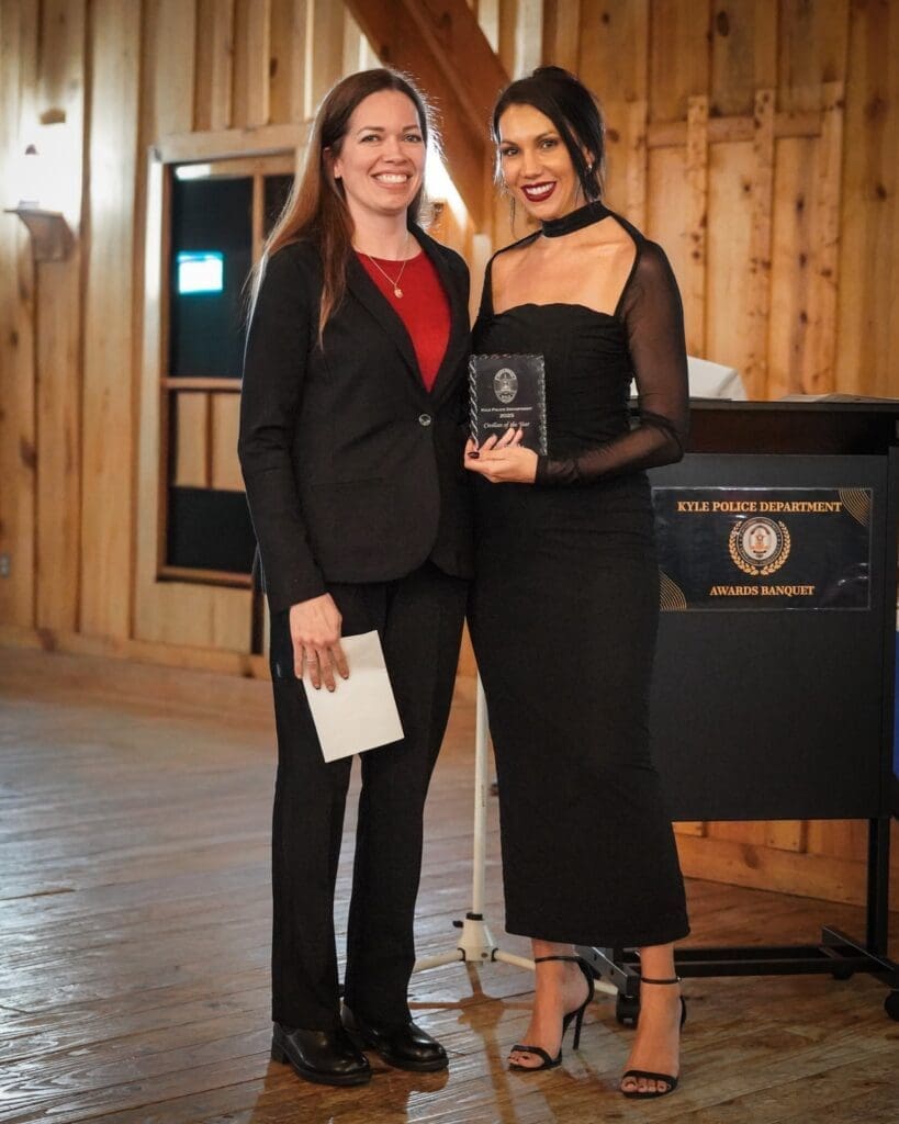 Two women stand indoors next to a podium labeled "Kyle Police Department Awards Banquet." One woman holds an award plaque and smiles, while the other stands beside her holding an envelope, both dressed formally.