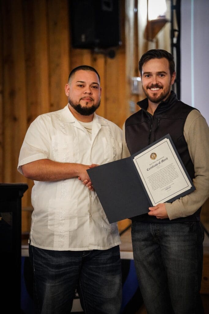 Two men stand indoors, shaking hands. One, in a white shirt, smiles at the camera. The other, in a dark vest, holds an open certificate of merit. Wood-paneled walls and a speaker are visible in the background.