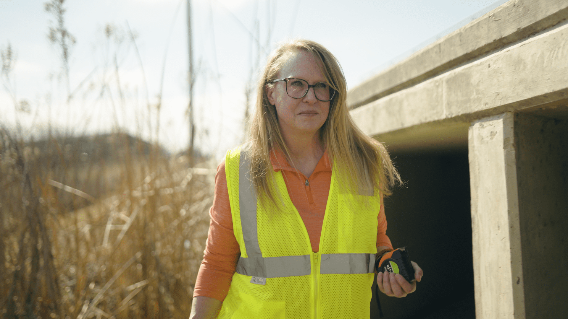 Kathy Roecker, Stormwater Program Manager is seen wearing glasses and a yellow safety vest stands outdoors near tall grasses and a concrete structure, holding an object in one hand, with sunlight illuminating the scene.