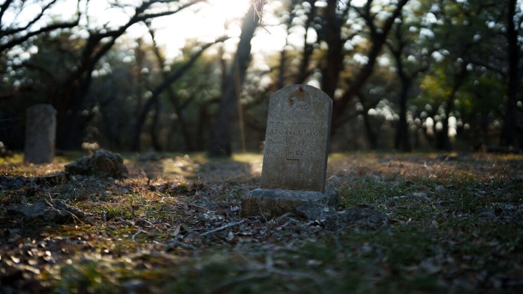 A weathered gravestone at the Kyle Family Cemetery stands in a sunlit, wooded cemetery with sunlight streaming through the trees and leaves scattered on the ground.