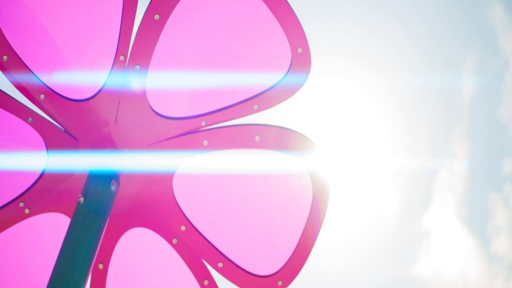 A close-up of a large, pink, flower-shaped structure with sunlight and lens flare creating blue streaks across the image, set against a bright sky.