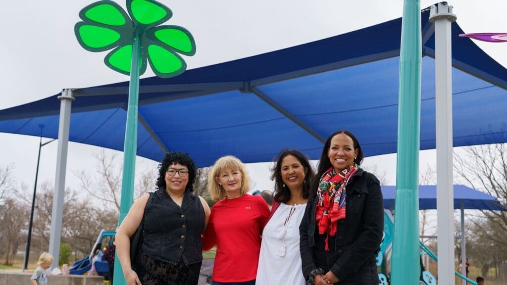 Four women stand together smiling at the Sky Blooms art installation ribbon cutting at Steeplechase Park.  Trees and playground equipment are visible in the background.