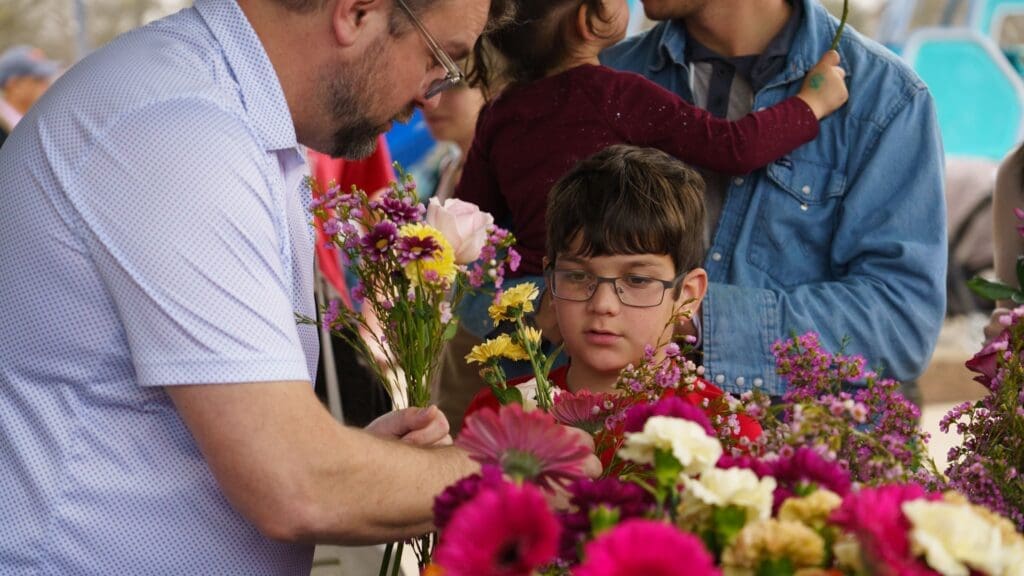 A man and a boy with glasses select flowers from a vibrant display at the Sky Blooms Art Installation ribbon cutting at Steeplechase Park. Another adult holds a child in the background, and colorful blooms fill the foreground.