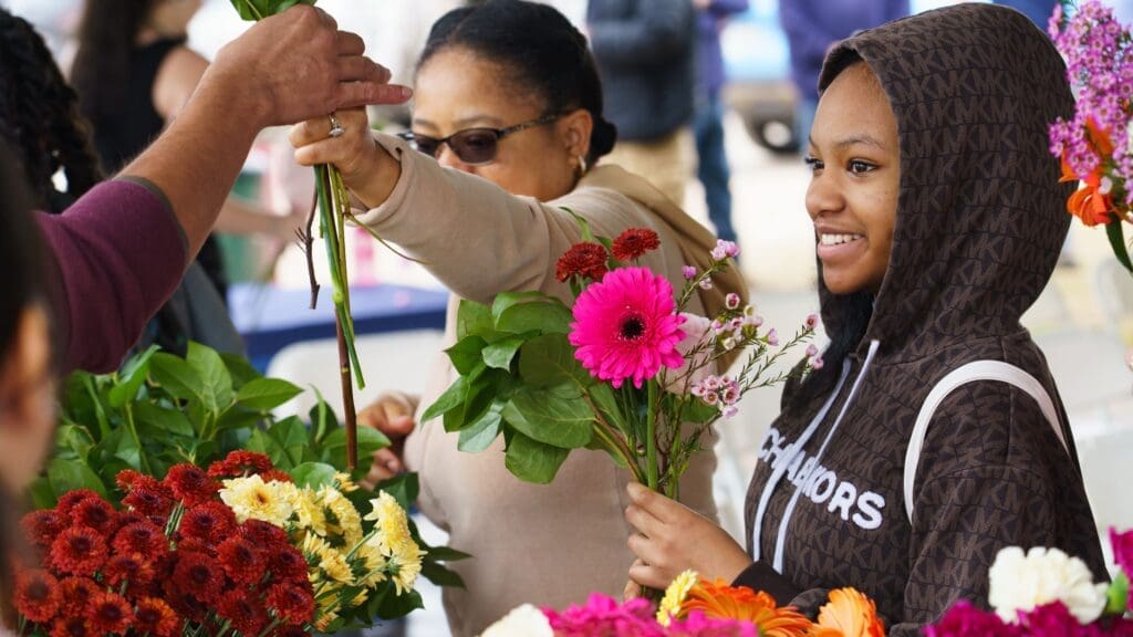 A smiling young woman in a brown hoodie holds a bouquet of vibrant flowers at the Sky Blooms Art Installation ribbon cutting at Steeplechase Park, while another woman picks out flowers from an assortment of colorful blooms.