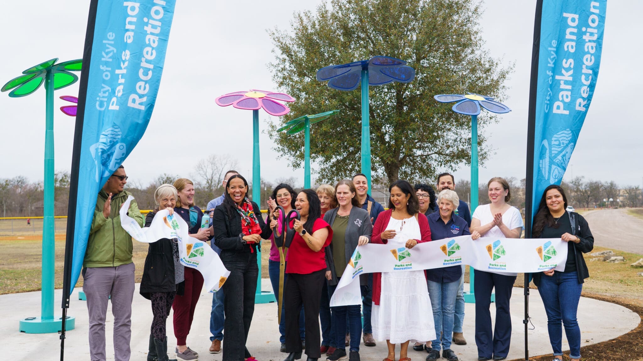 A group of people smile and hold a ribbon at an outdoor ribbon-cutting event near colorful flower sculptures and “Parks and Recreation” banners. Trees and a path are visible in the background.