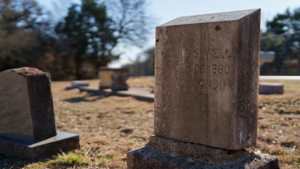 A weathered gravestone at Skyview Cemetery stands in the foreground of a sunlit cemetery, with other headstones and trees blurred in the background. The engraving on the stone appears faded with age.