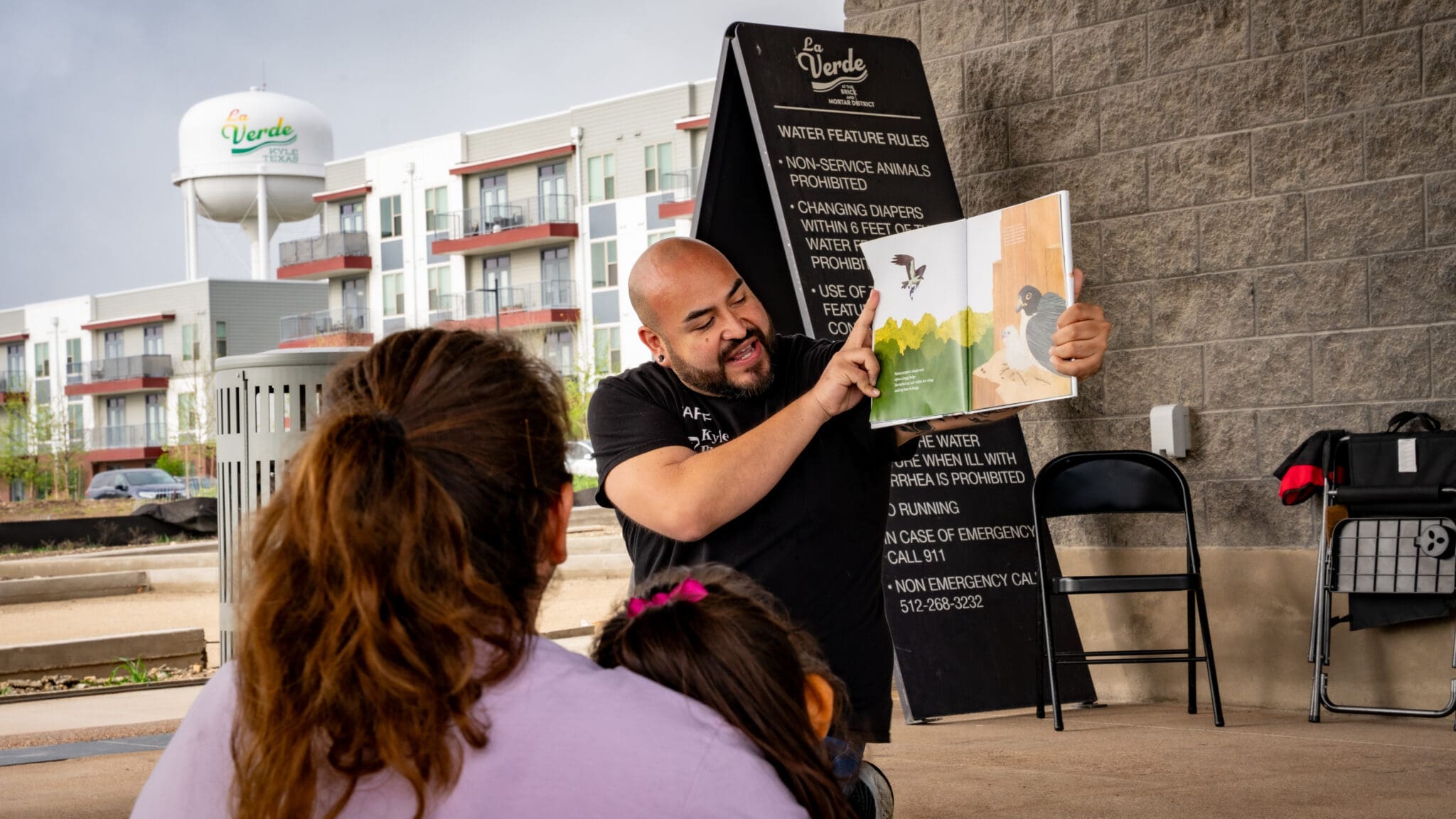 A man enthusiastically reads a picture book to two children outdoors, with buildings and a water tower in the background. A blackboard with rules and a folded chair are nearby.
