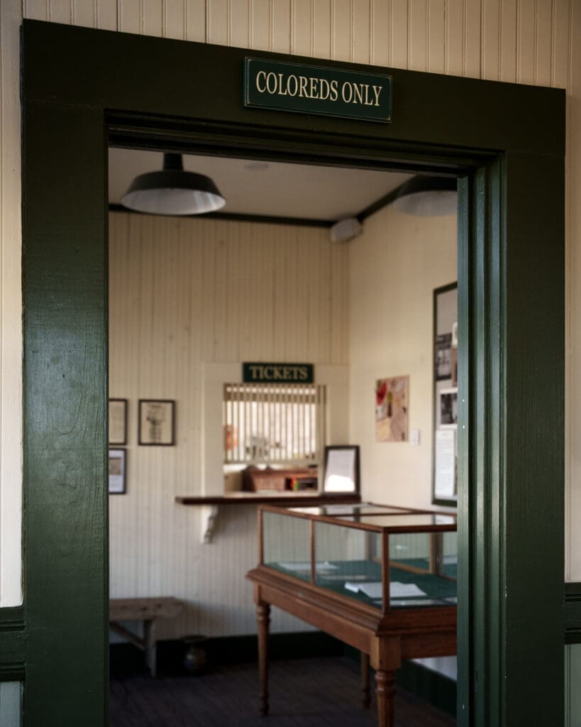A doorway with a sign above reading "COLOREDS ONLY" at the Kyle Train Depot leads into a room with a ticket window, a display case, and walls decorated with framed documents. The scene recalls segregation-era policies.