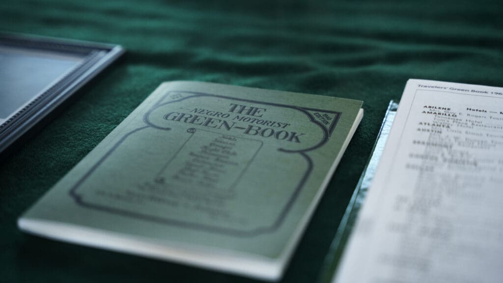 A vintage copy of "The Negro Motorist Green-Book" lies on a green surface next to a framed item and a sheet of paper with typed text at the Kyle Train Depot.