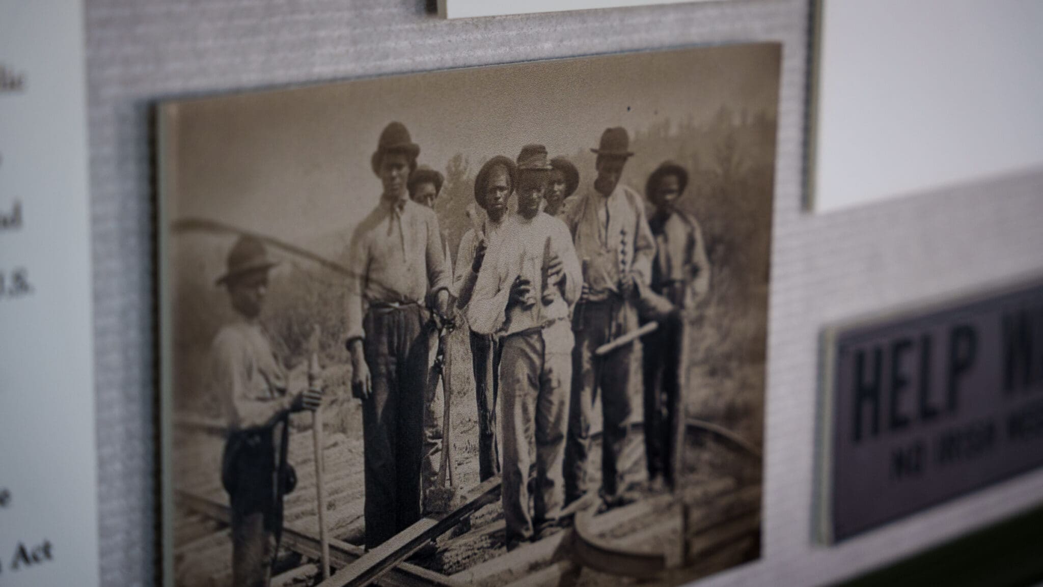 A sepia-toned historic photograph at the Kyle Train Depot shows a group of men standing on a railroad track, holding tools and wearing hats and work clothing, with blurred informational signs next to the photo.