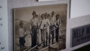 A sepia-toned historic photograph at the Kyle Train Depot shows a group of men standing on a railroad track, holding tools and wearing hats and work clothing, with blurred informational signs next to the photo.