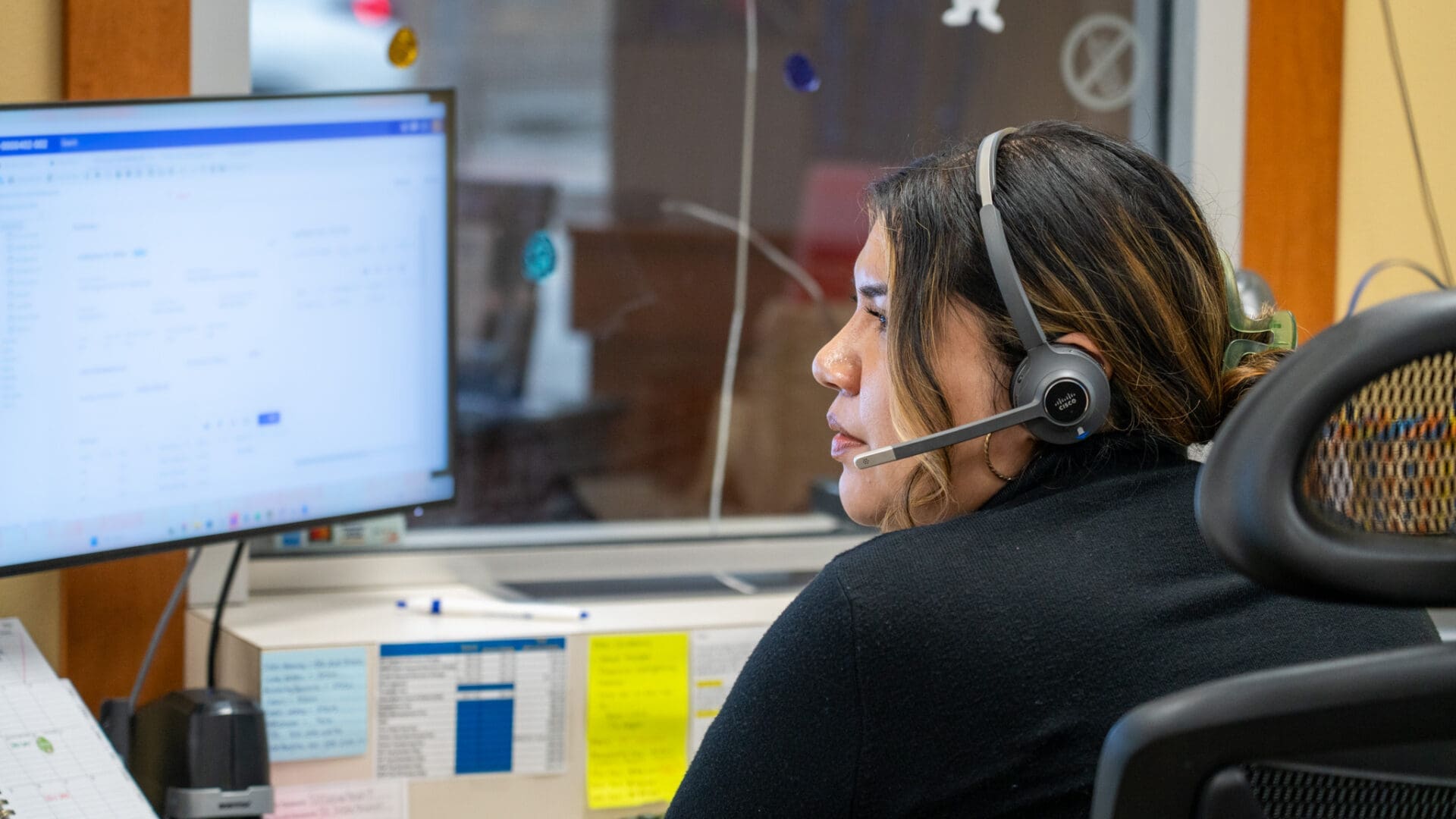 A woman wearing a headset sits at a desk and looks at a computer monitor with documents open. She is in an office setting with notes and papers visible around her workstation.
