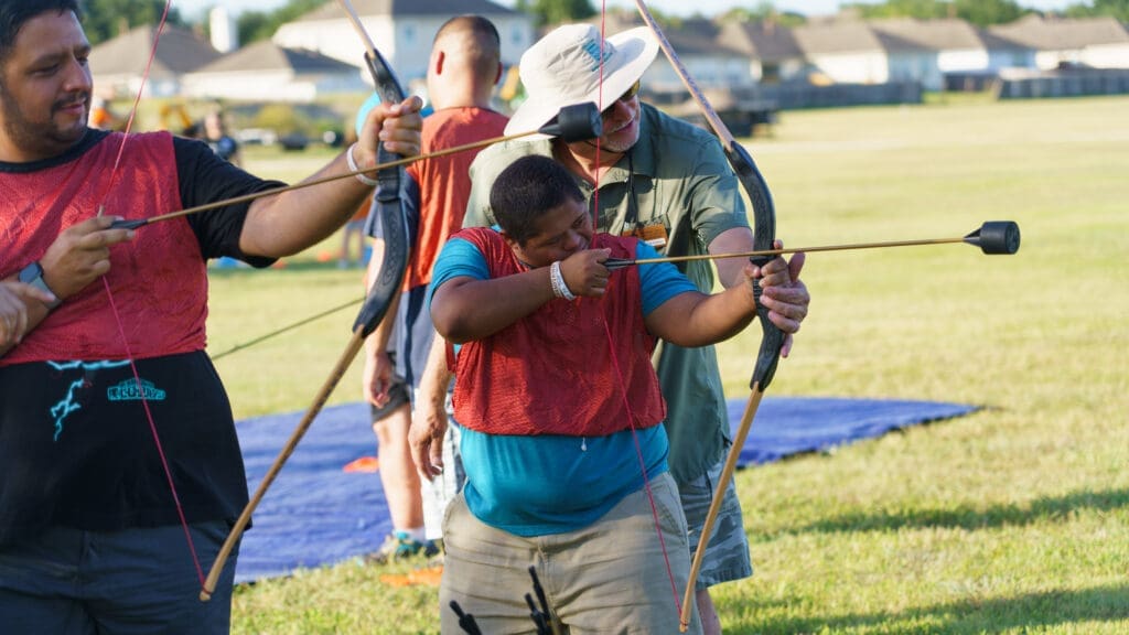 A group of people practice archery outdoors on grass, wearing red vests at the 2025 Adaptive Field Day. An instructor helps a participant aim a bow and arrow, while others stand nearby holding bows. Houses and trees are visible in the background.