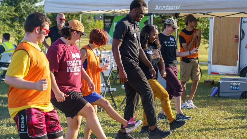 A group of people, some wearing orange vests, stand in a line outdoors, raising one leg as if participating in a group exercise or dance, with a tent and greenery visible in the background.