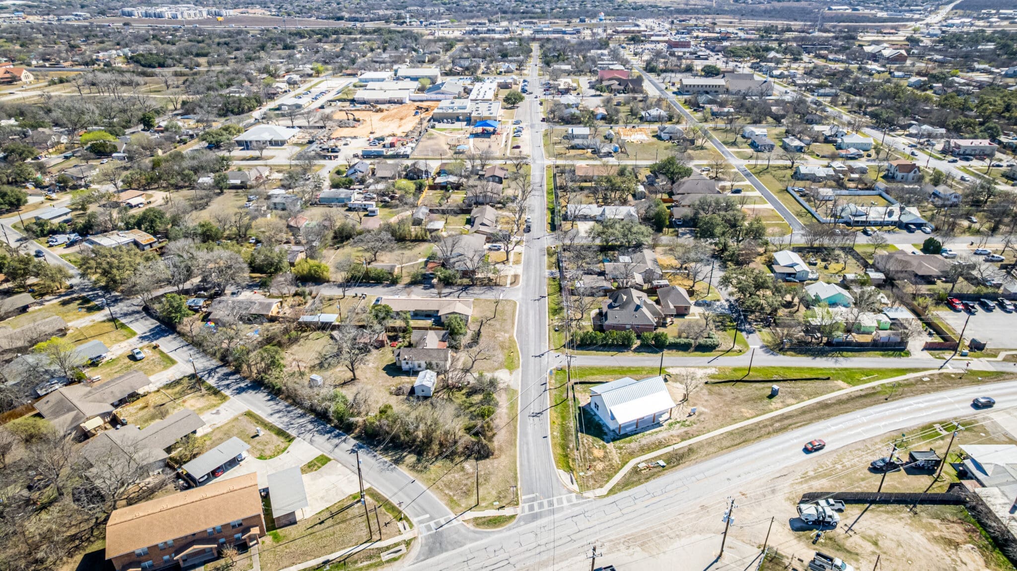 Aerial view of a suburban neighborhood with houses, trees, and streets intersecting. There are cars on the roads, a mix of residential and commercial buildings, and a large area under construction in the distance.