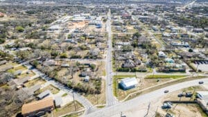 Aerial view of a suburban neighborhood with houses, trees, and streets intersecting. There are cars on the roads, a mix of residential and commercial buildings, and a large area under construction in the distance.