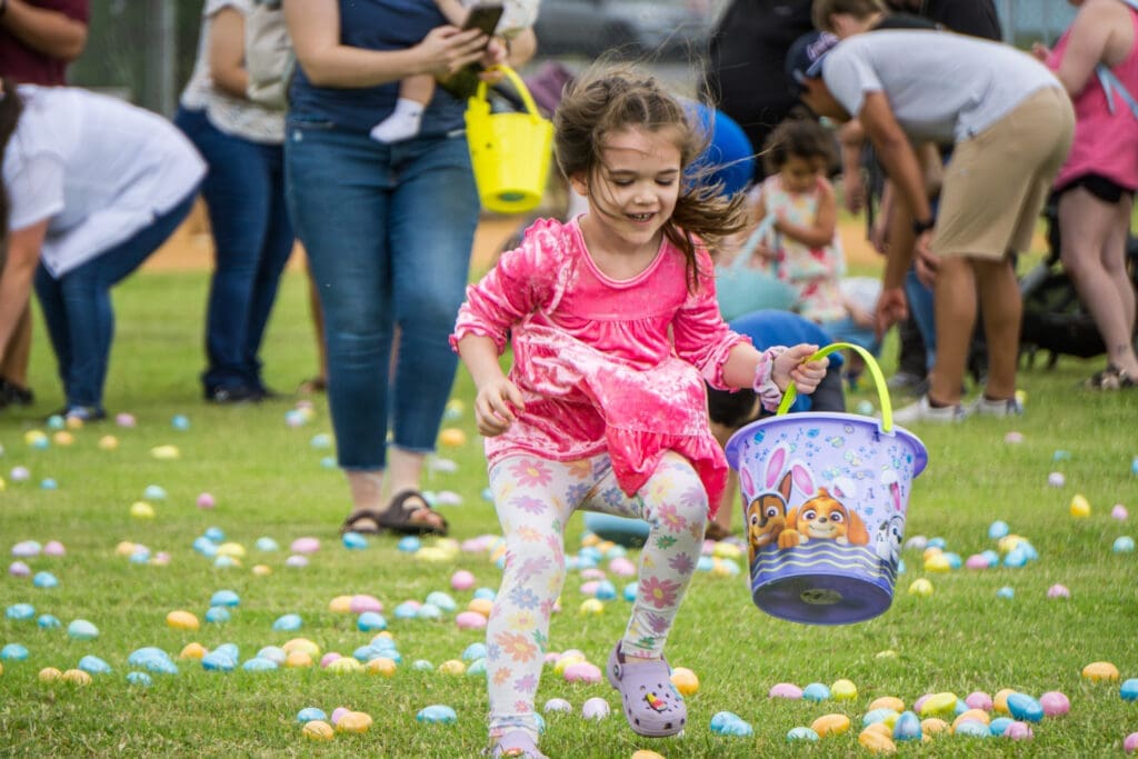 A young girl in a pink top runs excitedly on grass, holding an Easter-themed bucket, surrounded by colorful plastic eggs, while other children and adults search for eggs in the background.