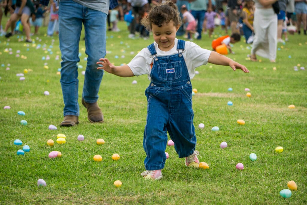 A young child in denim overalls smiles while walking on grass covered with colorful plastic Easter eggs, surrounded by people participating in an outdoor egg hunt.